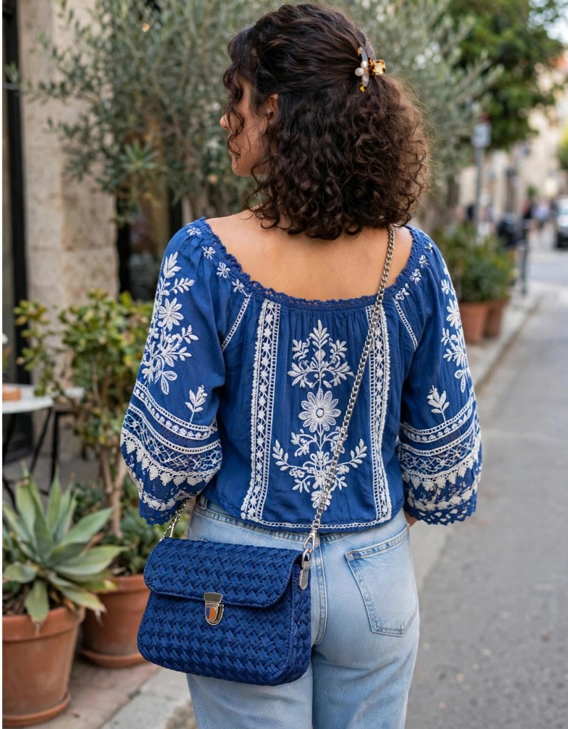 Donna vista da dietro che indossa una blusa blu ricamata, con capelli ricci e una borsa a tracolla blu. La scena si svolge in una strada di una città.